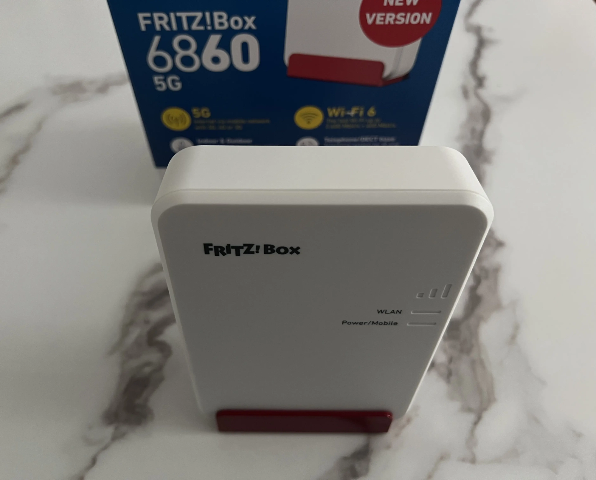 FRITZ!Box 6860 5G posée verticalement sur son support rouge devant sa boîte