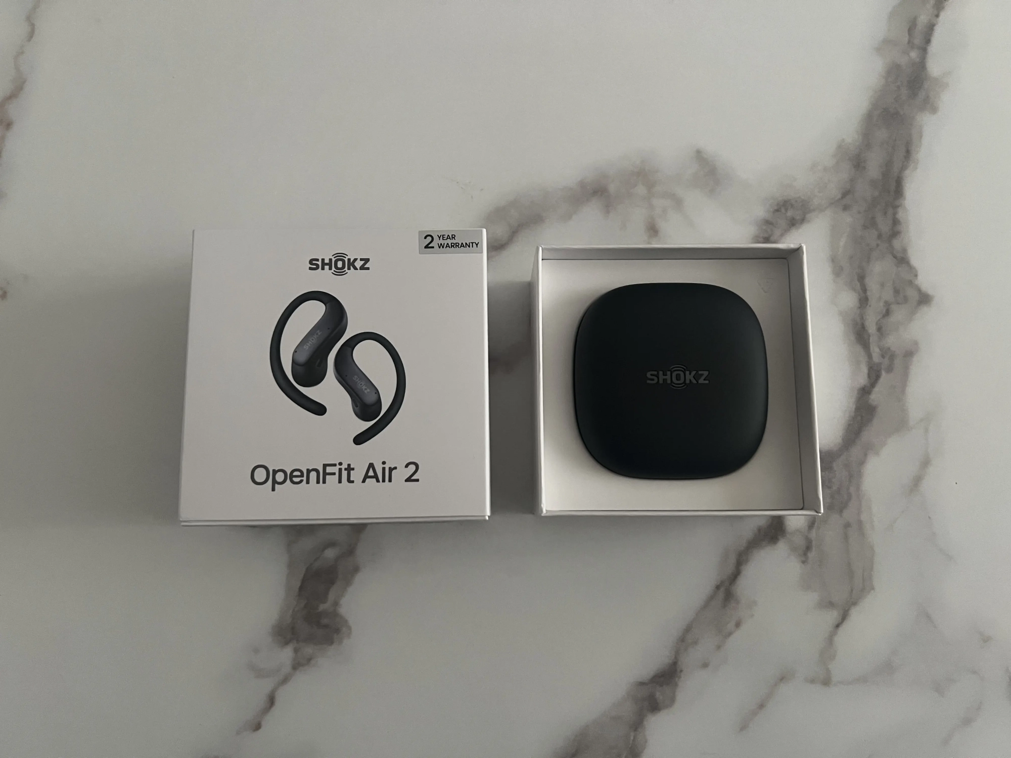 Boîte Shokz OpenFit Air 2 blanche à côté du boîtier de recharge noir dans son emballage